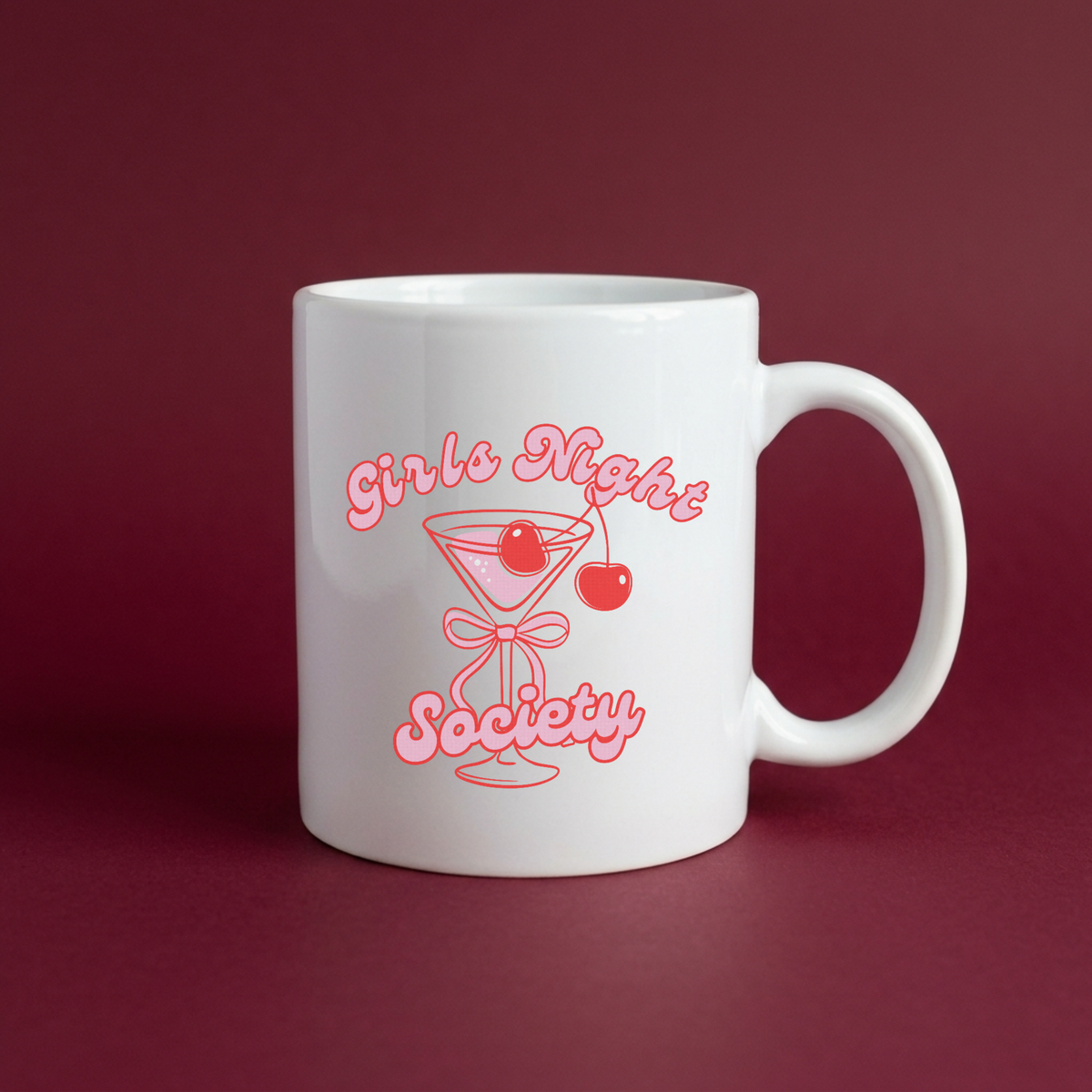 A white ceramic coffee mug with a pink graphic that reads 'Girls Night Society' around an illustration of a martini glass with a cherry, set against a dark burgundy background.