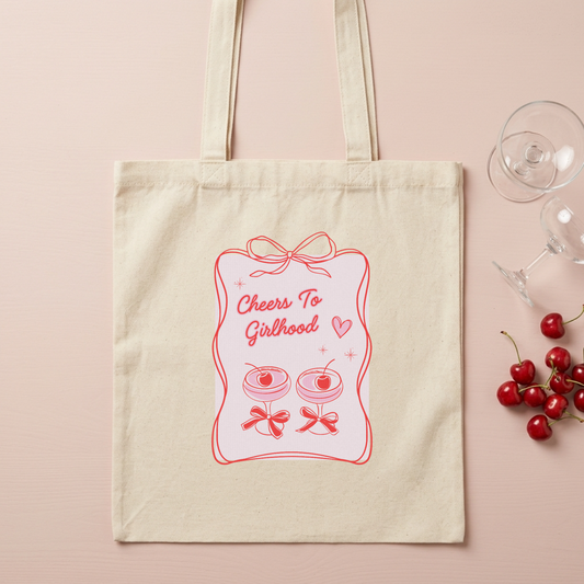 A cream canvas tote bag with a pink graphic that reads 'Cheers To Girlhood' with illustrations of macarons and cherries, displayed on a light pink background with cherry and wine glass props.